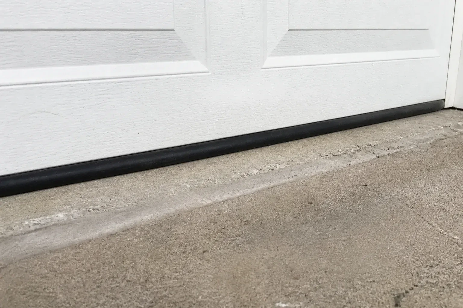 Black bottom seal along a closed white garage door at a concrete threshold. — Garage Door Bottom Seal Replacement, London