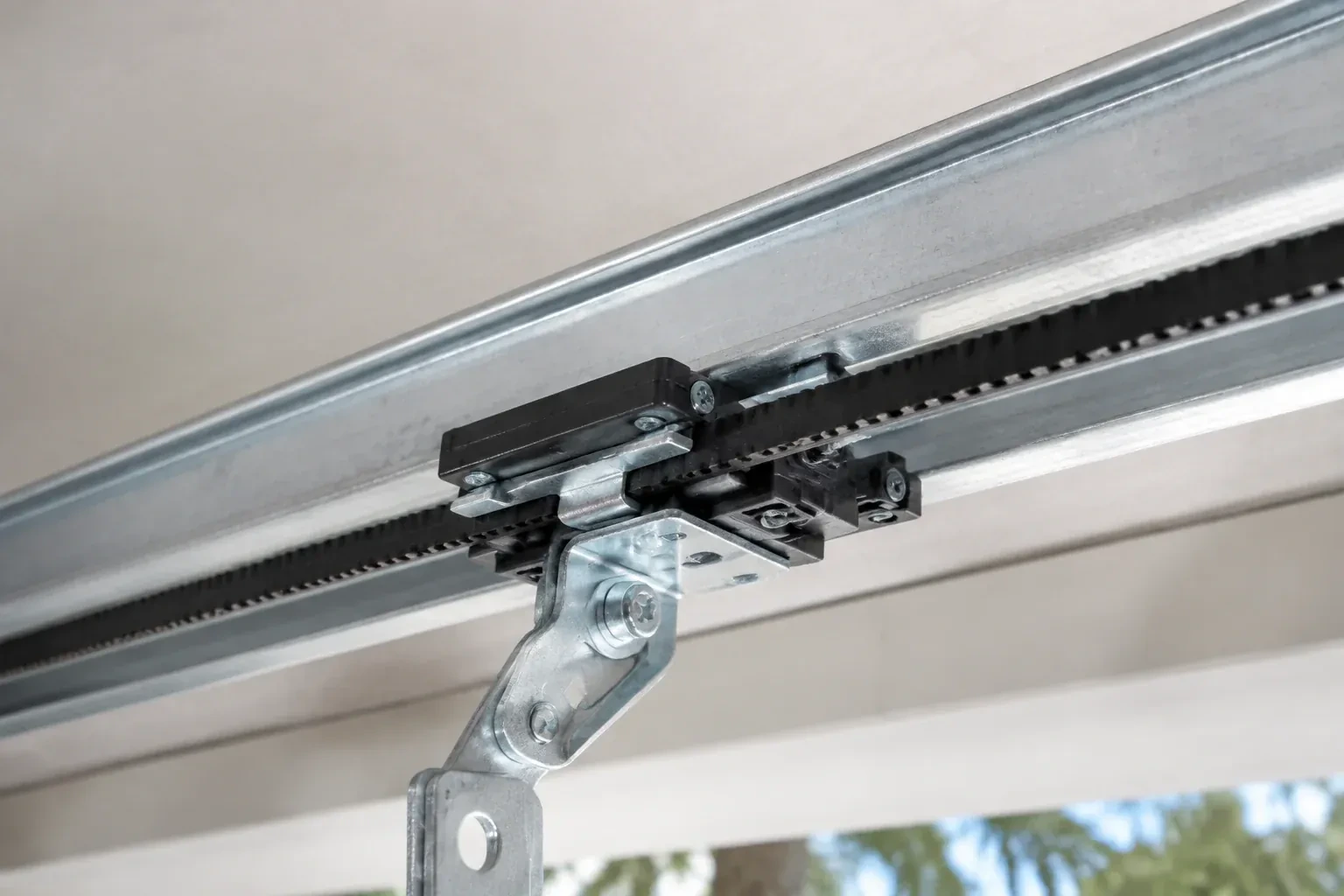 Black plastic trolley and bent galvanized bracket on a steel opener rail with a toothed black belt, against a ceiling. — Belt-Drive Opener Repair, London